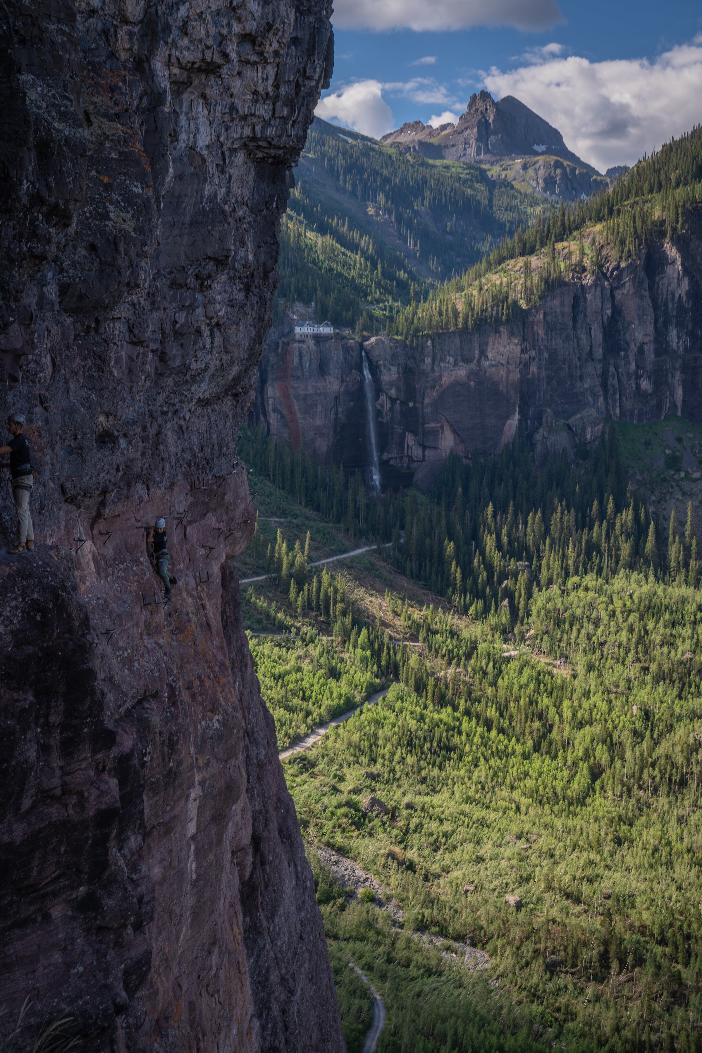 Cliffside with climber on the left with green trees below. Bridal veil falls waterfall and pump house above with mountain peak beyond