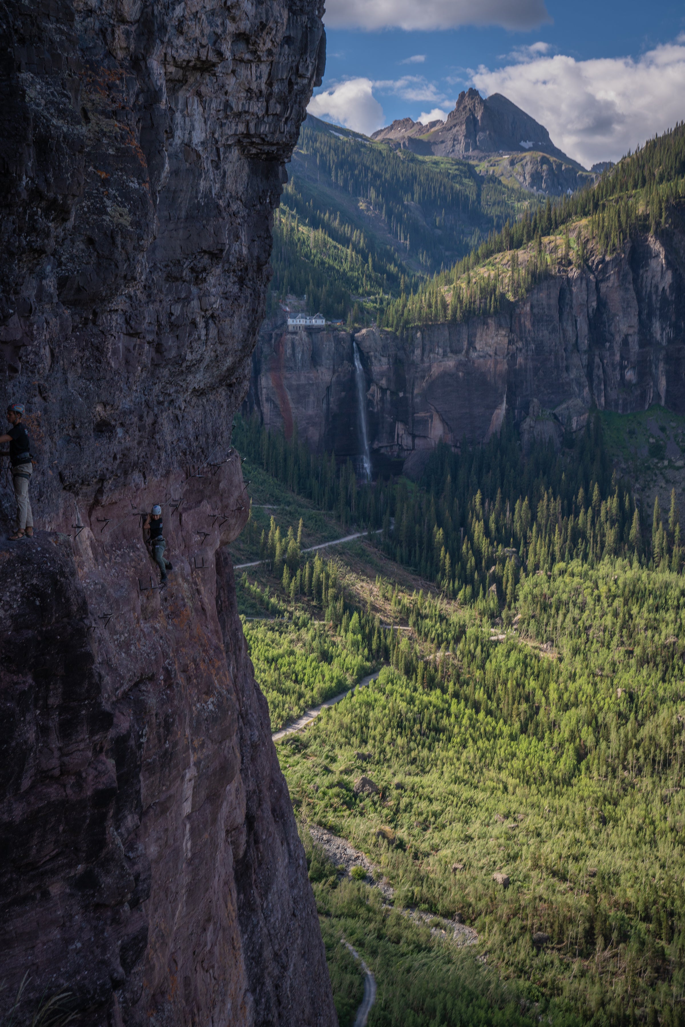 Cliffside with climber on the left with green trees below. Bridal veil falls waterfall and pump house above with mountain peak beyond 