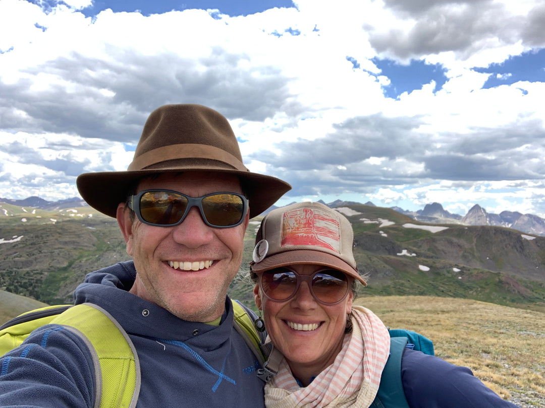 Tim Lafferty in a brown hat with and sunglasses, Jenna Cichanski with a brown hat and sunglasses, both in backpacks, with a cloudy sky and mountainous background