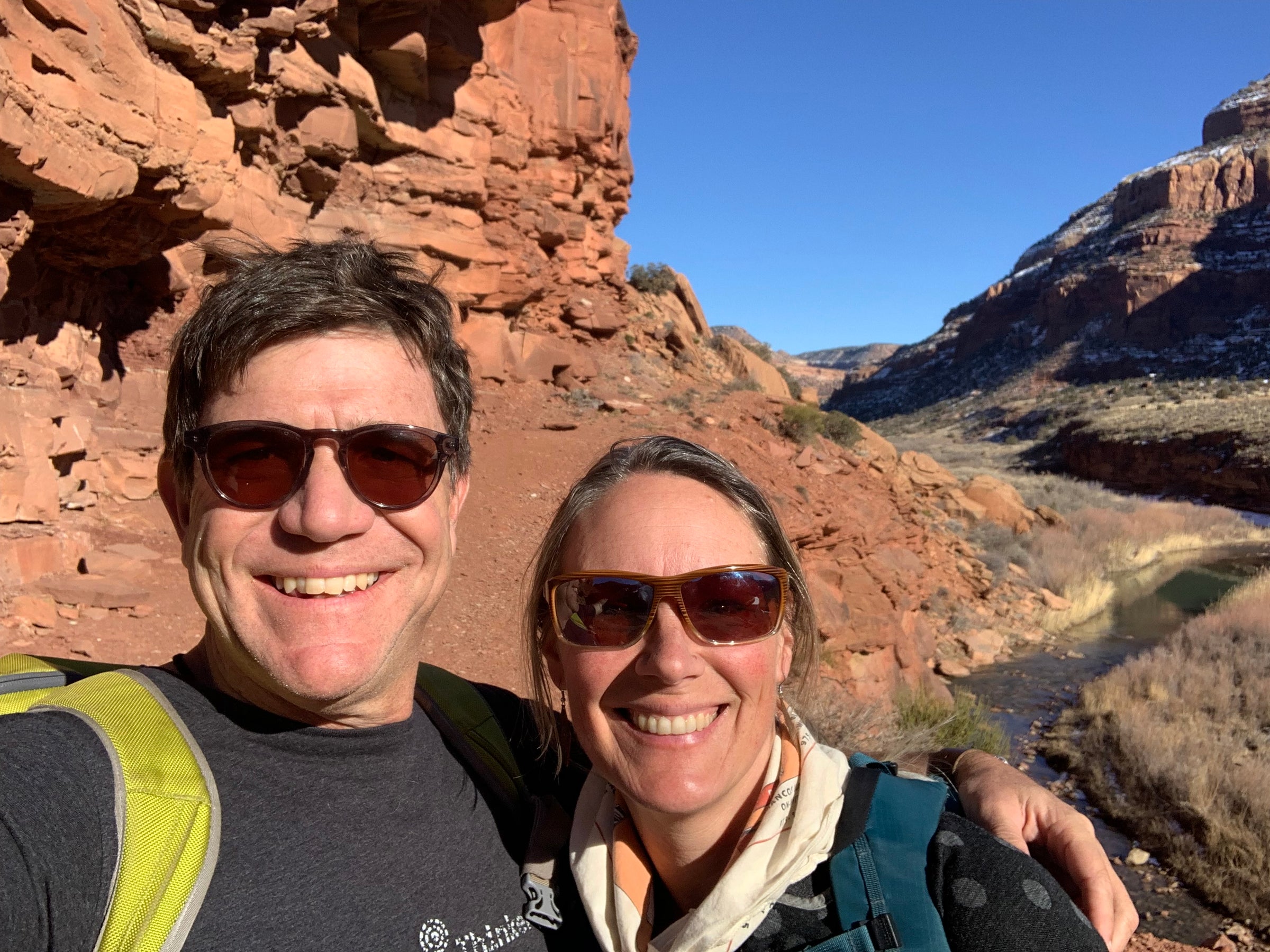 Tim Lafferty wearing a black t-shirt and brown sunglasses, Jenna Cichanski wearing a multi-colored sscarf and sunglasses, on a rocky hike with a creek running through a dry field