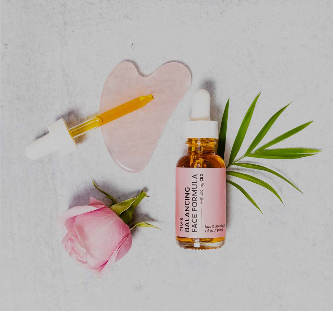 Pink rose with rose quartz gua sha and Tim's Balancing Face Formula glass bottle with a leaf on a white background