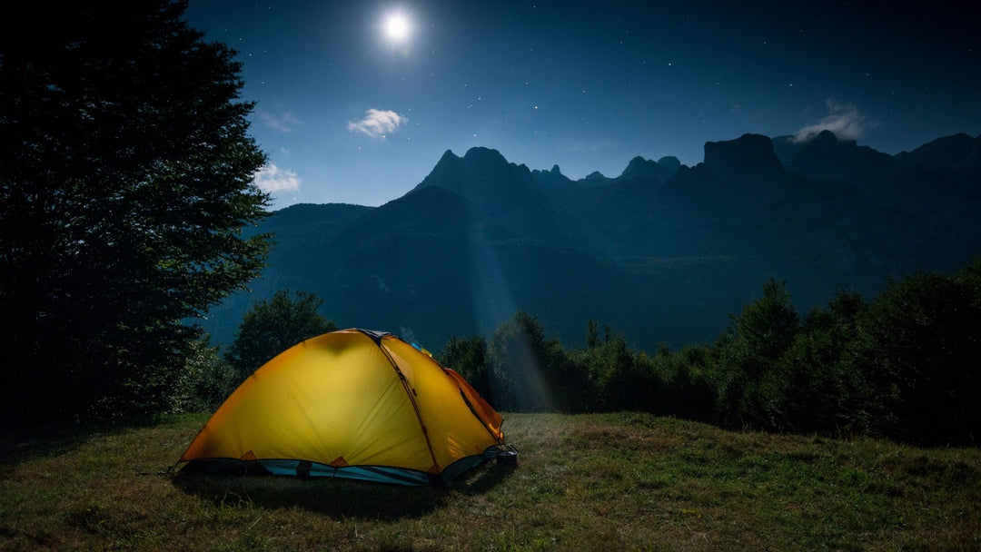 Glowing tent on a grassy field with trees, jagged mountains and the moon glowing 