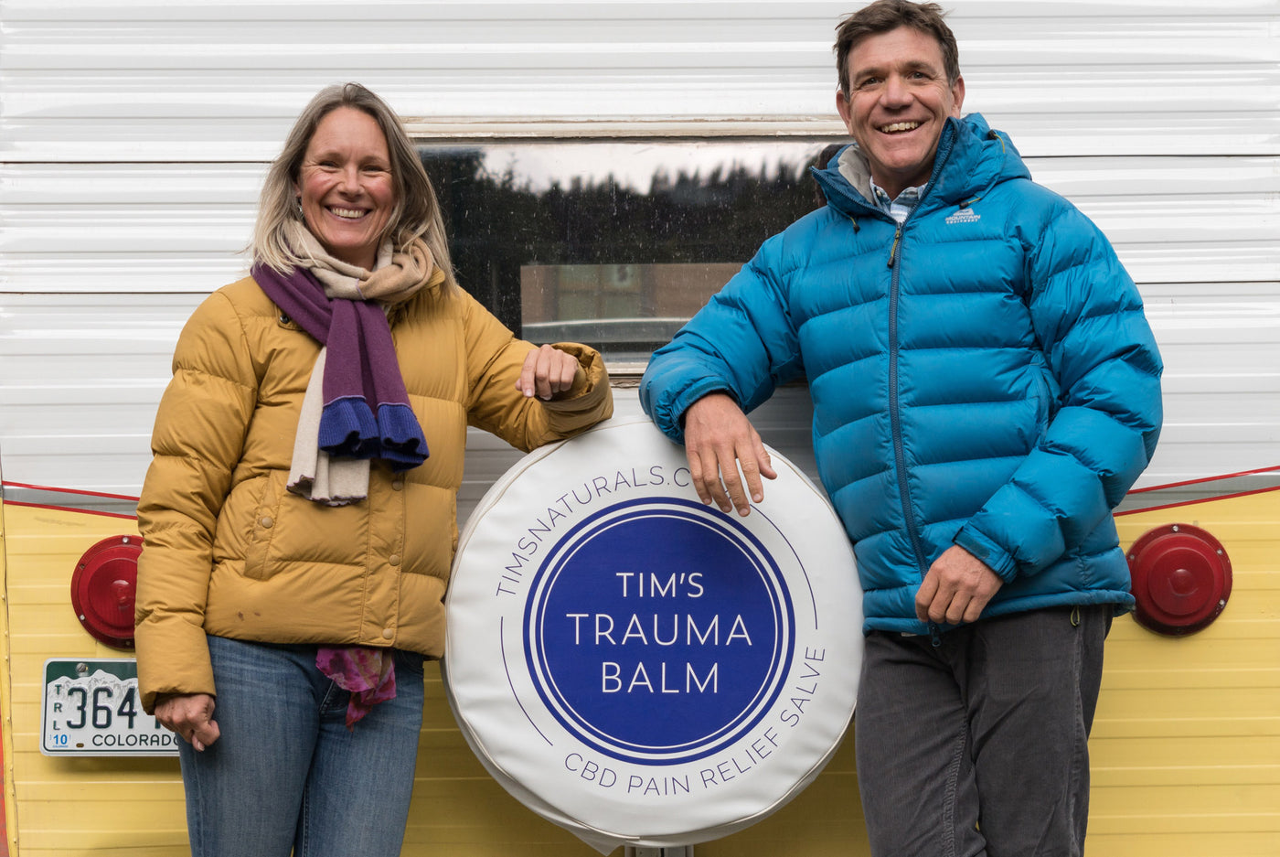 Jenna Cichanski in a mustard yellow puffy jacket, purple, blue and ivory scarf, and jeans, Tim Lafferty in bright blue puffy jacket, plaid button down, grey pants, in front of a shasta white and yellow camper, with the Tim's Trauma Balm blue circle