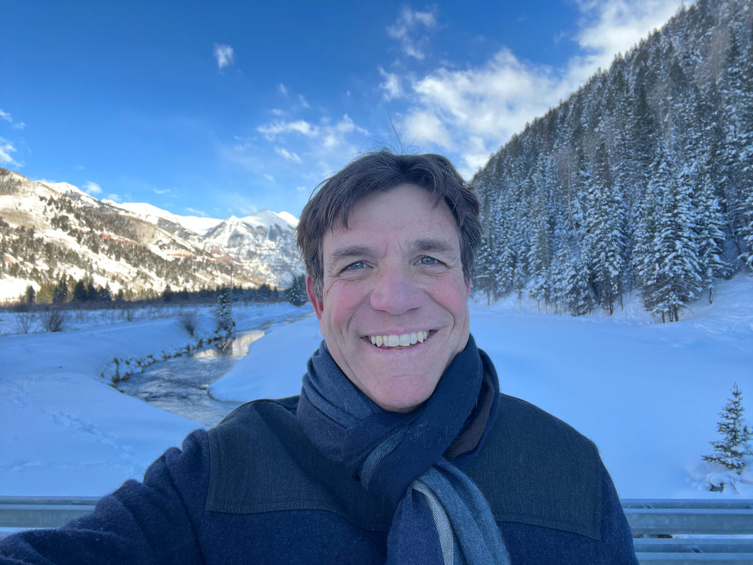 Tim Lafferty wearing a grey coat and grey and blue scarf, smiling at the camera. Snowy Telluride valley floor with San Miguel river running through and Ajax peak in the background with pine and spruce trees in between
