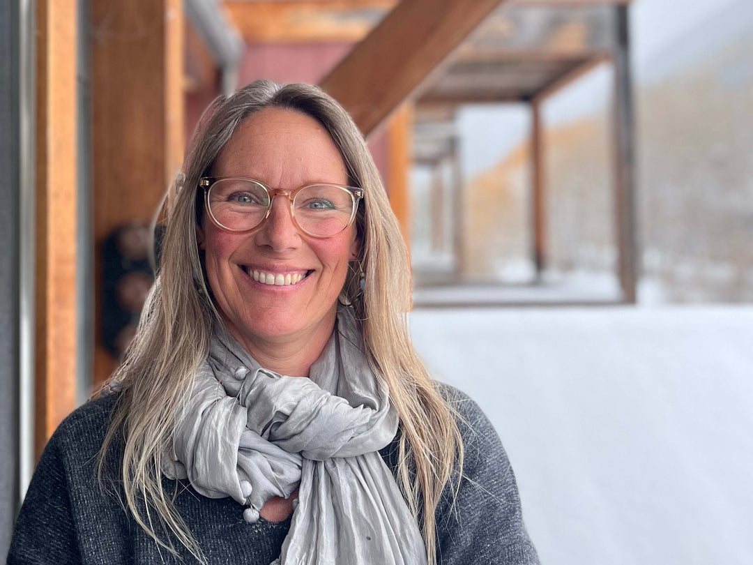 Jenna Lafferty with long blonde hair, glasses, smile and light grey scarf, smiling in front of a wooden beam with snow on the ground