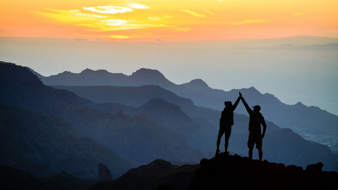 Two people high fiving on a hike with mountains and a colorful sunset in the background