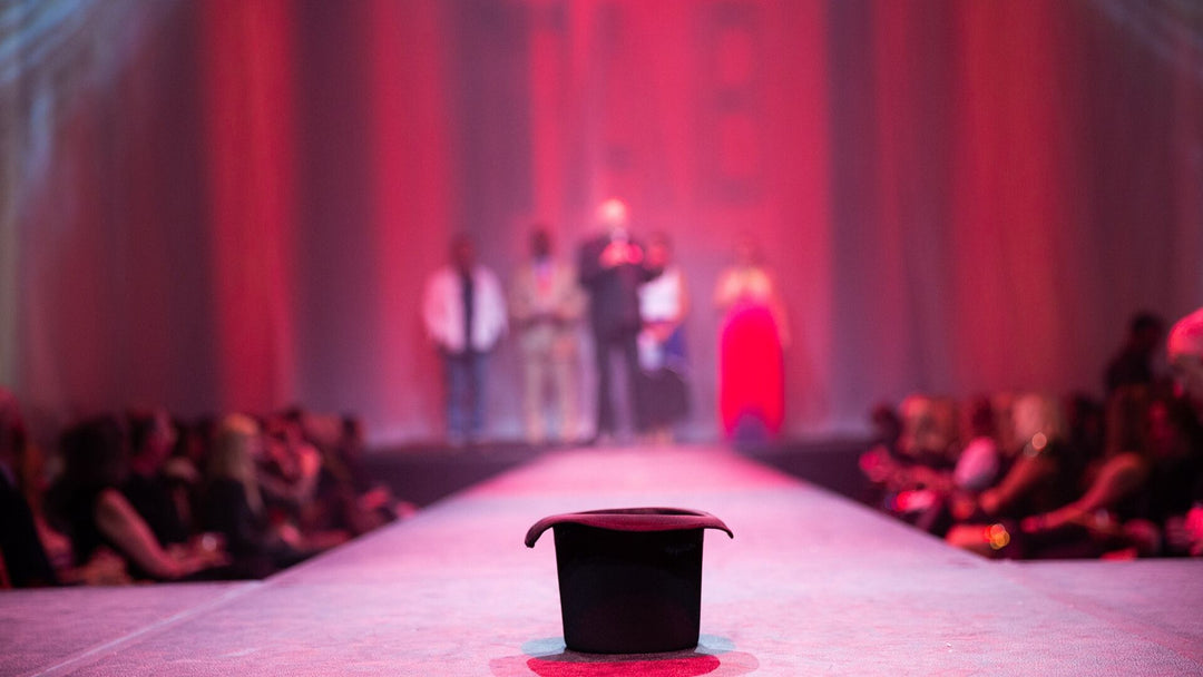 Fashion runway with top hat sitting center stage and audience on either side. Models blurred in the background with pink curtain background