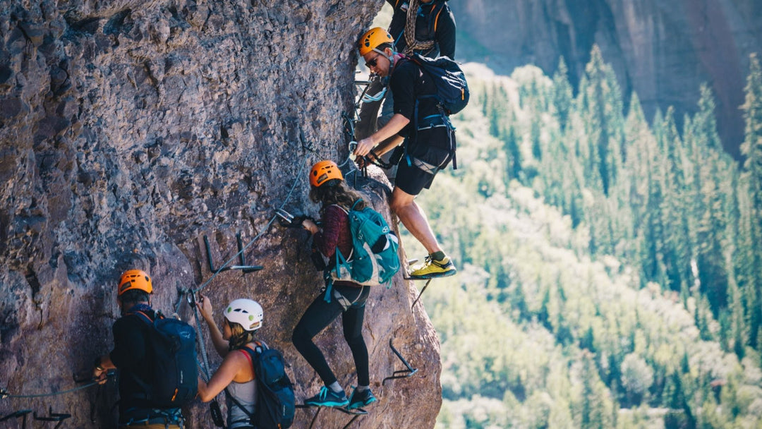 Five people mountaineering with rock climbing helmets and harnesses on a cliffside with evergreen trees beyond