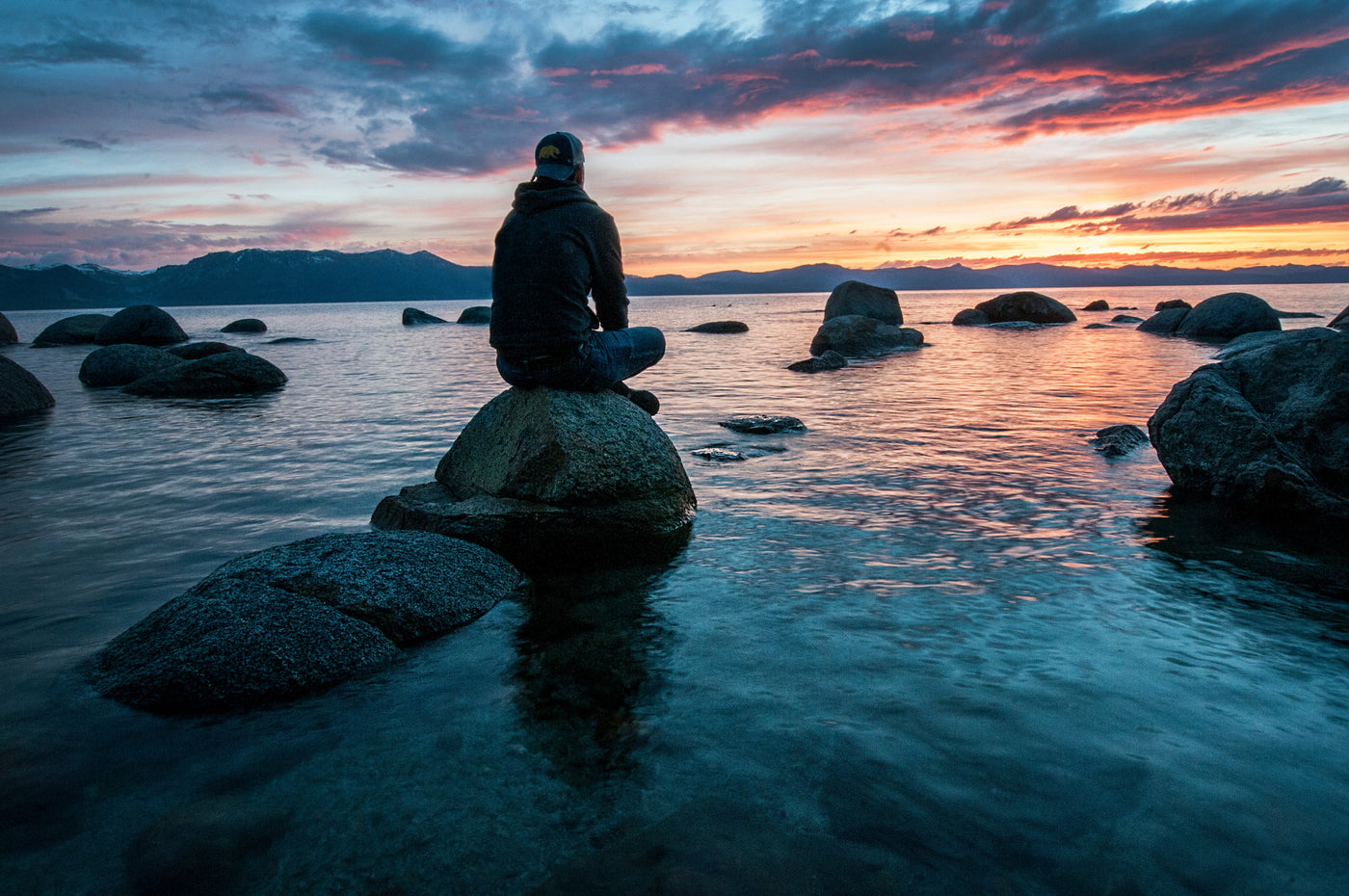 person sitting on a rock, surrounded by water, and other rocks. Mountains in the background, and colorful sunset with pink lined clouds