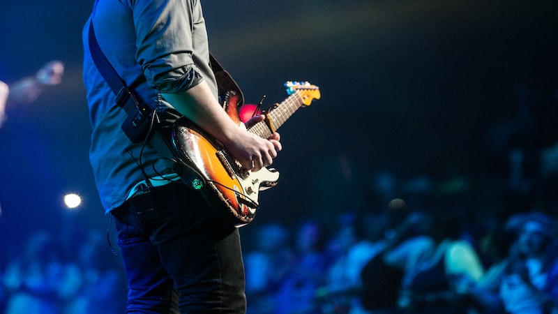 Guitarist from behind with electric guitar, grey shirt with rolled up sleeve and audience blurred beyond him