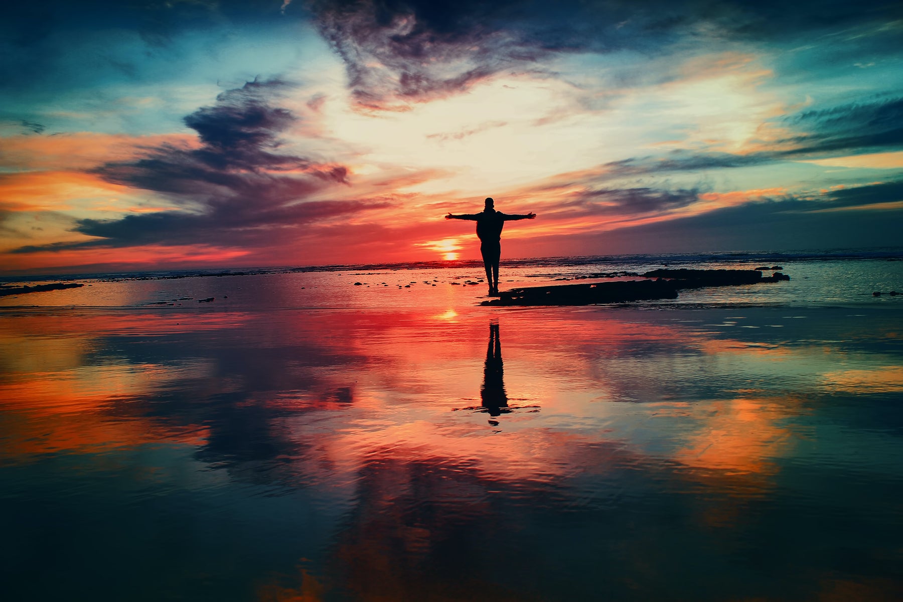 Colorful sunset reflecting on wet sand with a person's silhouette and arms outstretched