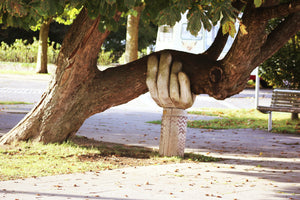 Wooden hand statue holding up a leaning tree with a park bench beyond